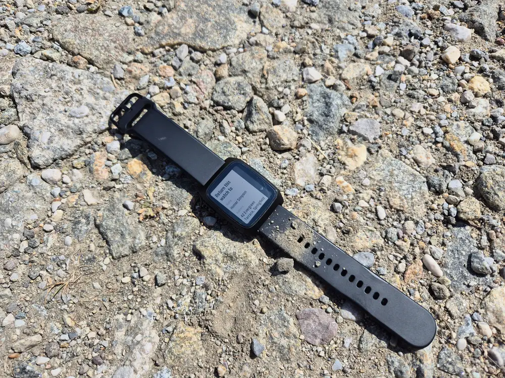 A Pebble watch lying on gravel where it was dropped, its screen showing the Return to contact card